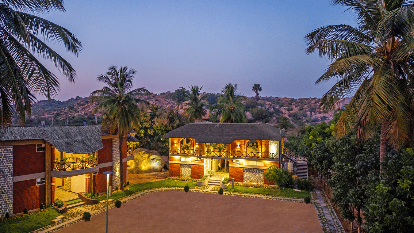 The resort buildings illuminated at dusk, showcasing the red-brick architecture and sandy courtyard - Nature Trails Ashoka Resort Hampi