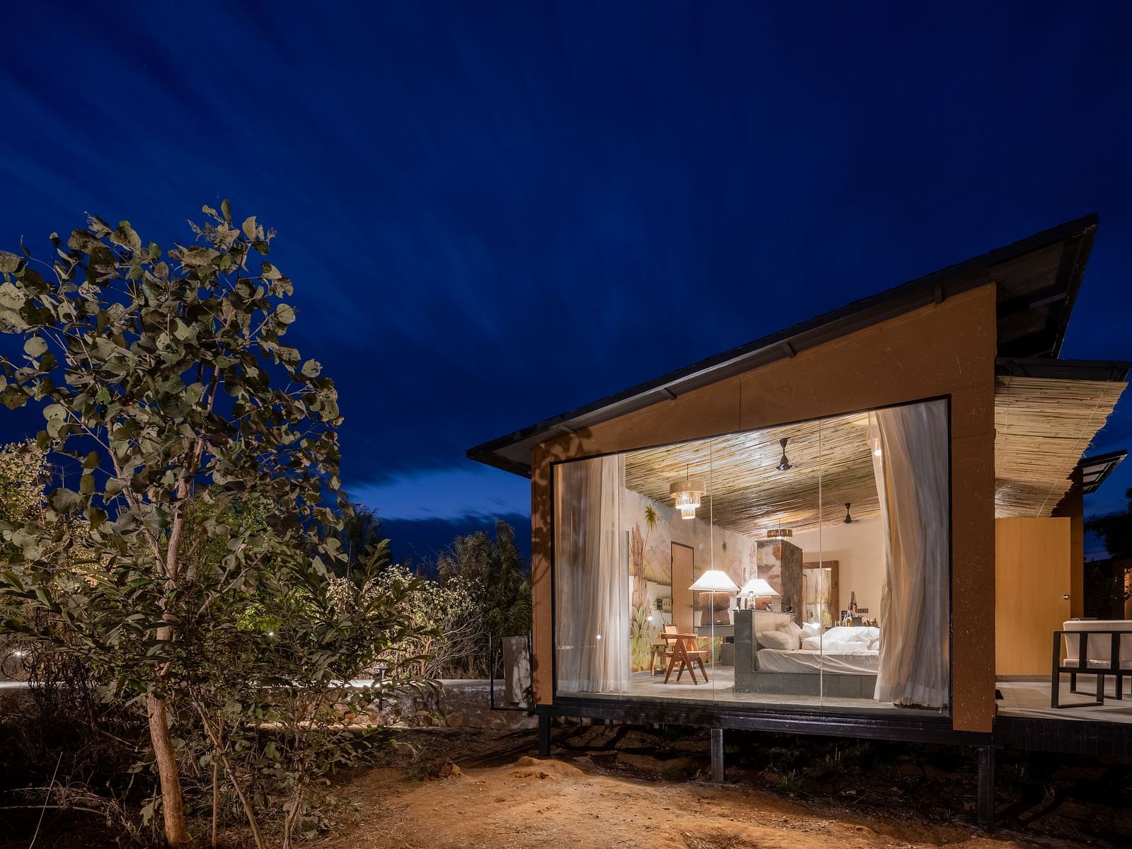 A facade view of a room with bright lighting and a tree outside it as seen in the night - Utsav Camp Sariska