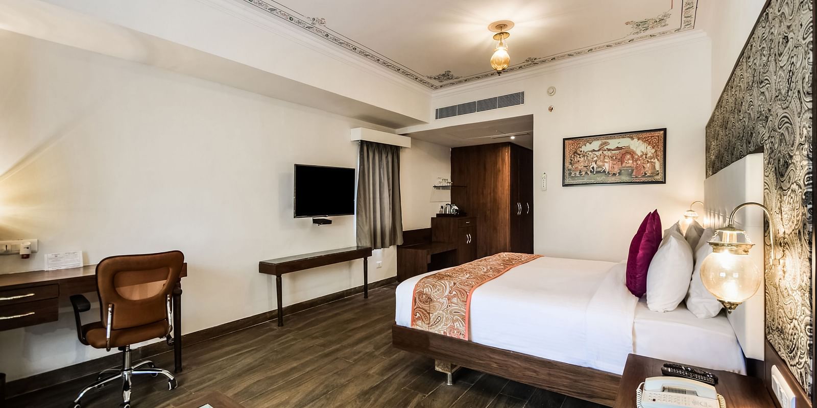 A spacious hotel room with a bed, desk, chair, and TV. - Bamboo Saa Resort & Spa, Udaipur