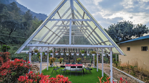 A white-framed, transparent-roofed gazebo or greenhouse structure with a green lawn inside set for dining, surrounded by red and green plants, with misty mountains in the background under a cloudy sky.