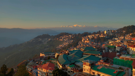 Scenic view of Darjeeling town with colourful hillside buildings, green rooftops, and winding roads, set against misty hills and snow-capped Himalayan peaks under a clear sky at sunrise.