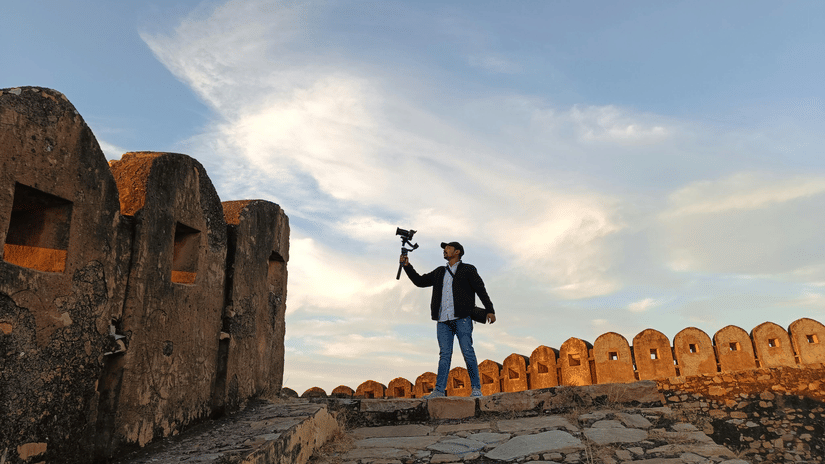 A man stands on the stone ramparts of a fort, holding a camera gimbal against a backdrop of soft evening clouds.