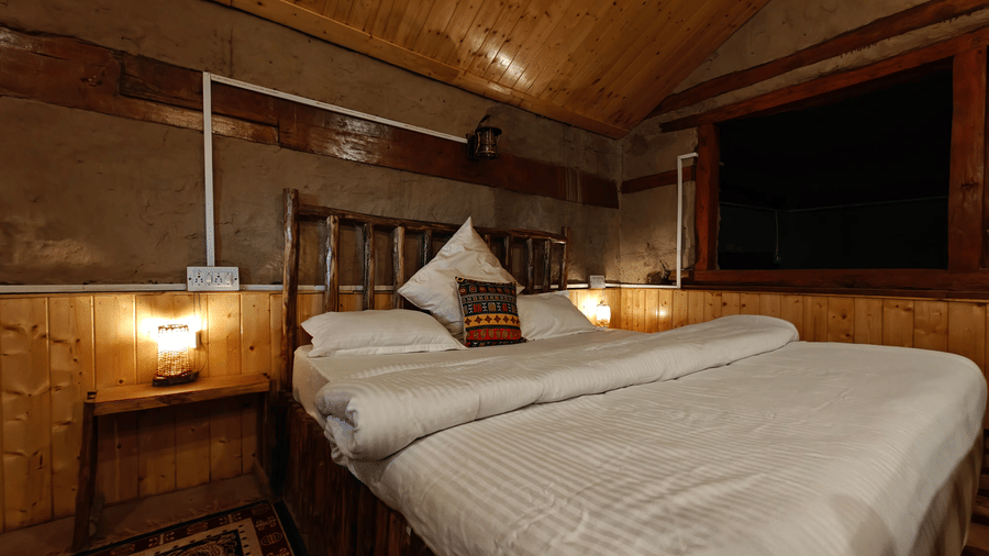 Corner view of a plush double bed at the Friendship Peak Cottage featuring 2 brightly-lit table on either side of the bed at Amara Upepo - The Sky Village, Manali.