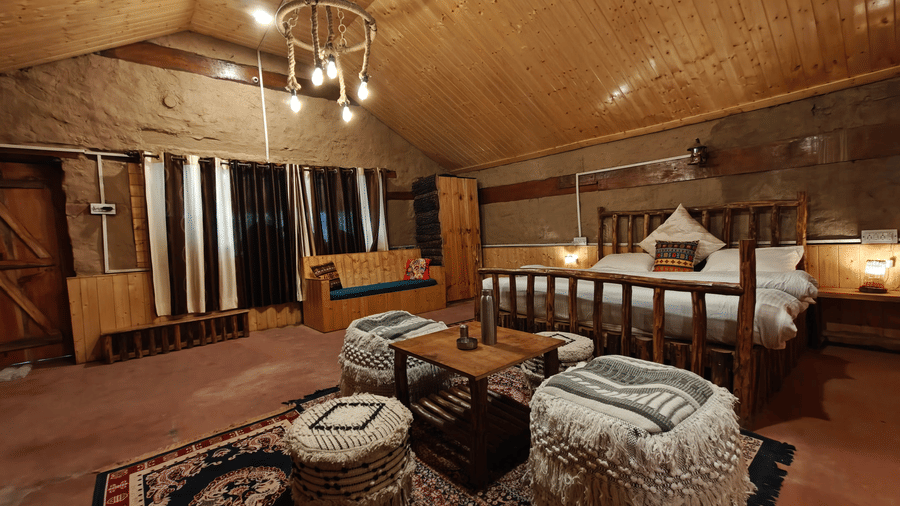 A wide-angle shot of the Friendship Peak Cottage with cosy wooden interiors featuring a double bed with a wooden bed frame with a common seating are in front of it at Amara Upepo - The Sky Village, Manali.