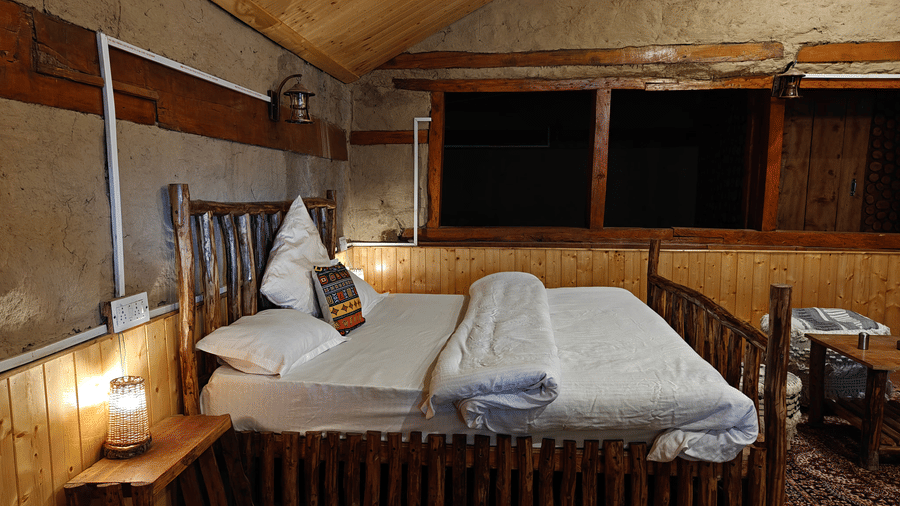 Side view of the double bed at the Friendship Peak Cottage featuring a bed side table with a small lamp kept on it at Amara Upepo - The Sky Village, Manali.