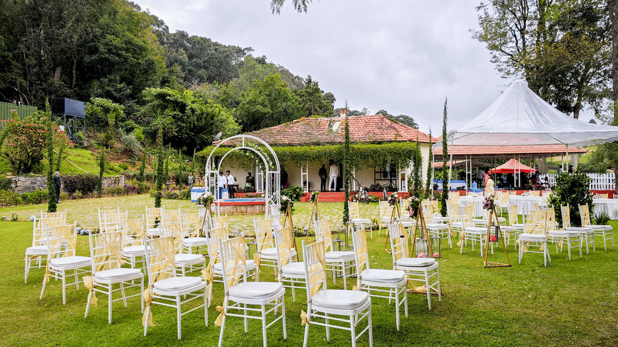 n outdoor event setting with a floral archway and a seating area with white chairs and yellow sashes on a green lawn, surrounded by lush foliage and trees.