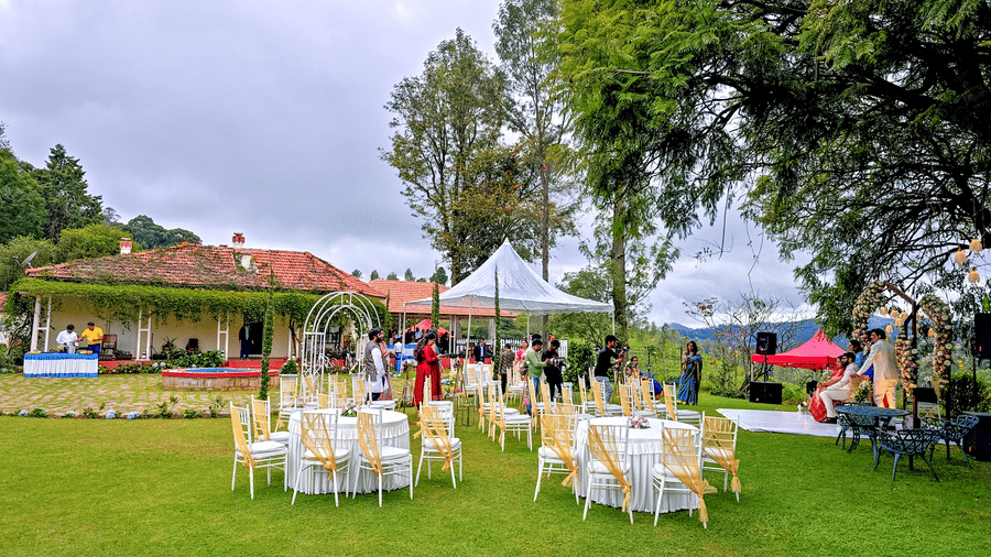 n outdoor event setting with a floral archway and a seating area with white chairs and yellow sashes on a green lawn, surrounded by lush foliage and trees.