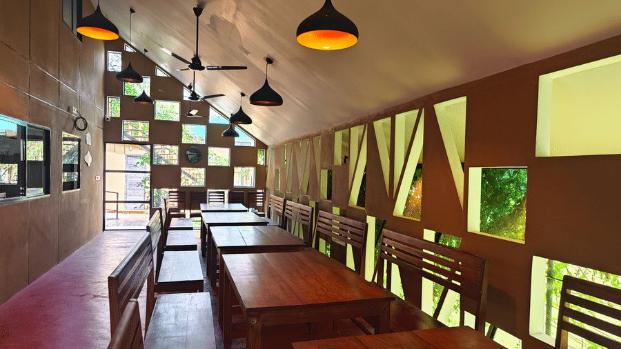 Interior of a rustic dining hall with long wooden tables, warm pendant lighting, and a unique geometric window wall - Ibex Resorts, Malampuzha (Kava Eco Camp and Caravan Park)
