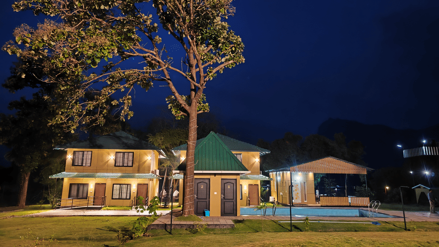 A warm night shot of the resort buildings illuminated against a dark blue sky with a tall tree in the foreground - Ibex Resorts, Malampuzha (Kava Eco Camp and Caravan Park)
