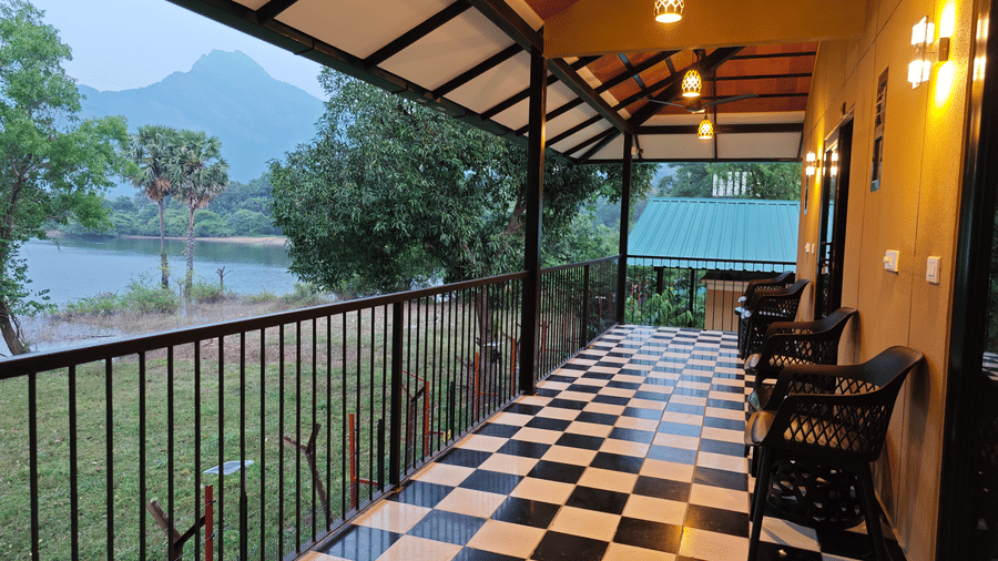A long veranda with black and white checker flooring and seating, offering a view of mountains and a lake - Ibex Resorts, Malampuzha (Kava Eco Camp and Caravan Park)