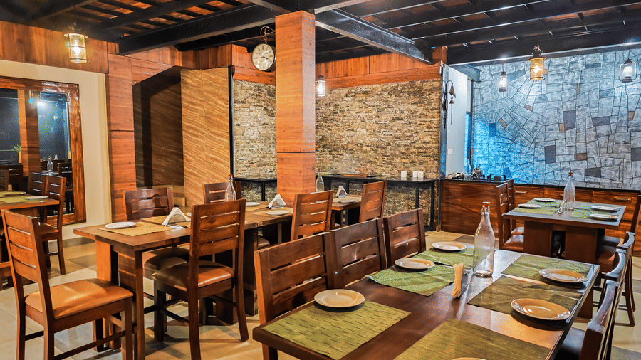 Rustic indoor dining space at Ibex Resorts Valparai Tale Rock featuring solid wooden tables brick accent walls warm pendant lighting and neatly arranged place settings creating a cosy and inviting atmosphere in the hills.