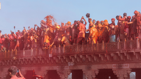 Crowd of people celebrating Holi on a temple balcony and street, throwing vibrant coloured powders, capturing the joyful festival atmosphere under a clear blue sky.