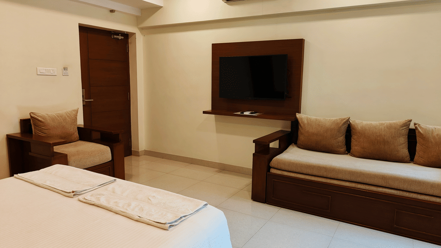 A well-lit room, featuring a large bed with white linens, a wall-mounted TV, and a sofa with brown cushions | A & M Rooms and Residences
