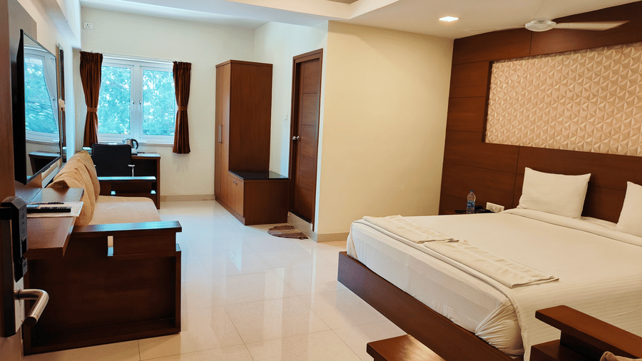 A spacious room featuring a large bed with white linens, a desk area near a window, and dark wooden furniture on a reflective light-coloured floor | A & M Rooms and Residences