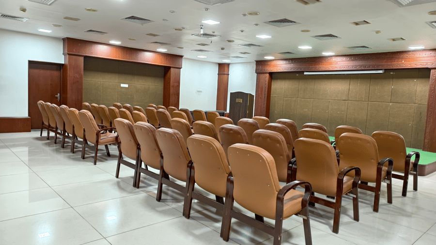 An indoor conference hall showing rows of seating, wooden podium, and stage setup beneath ceiling light fixtures at DSF Grand Plaza, Tuticorin.