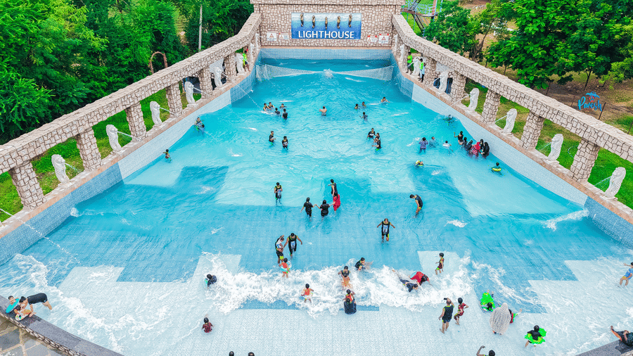Large outdoor swimming pool with people in the pool, bordered by steps and greenery all around - Lighthouse Waterpark & Resort, Nagpur