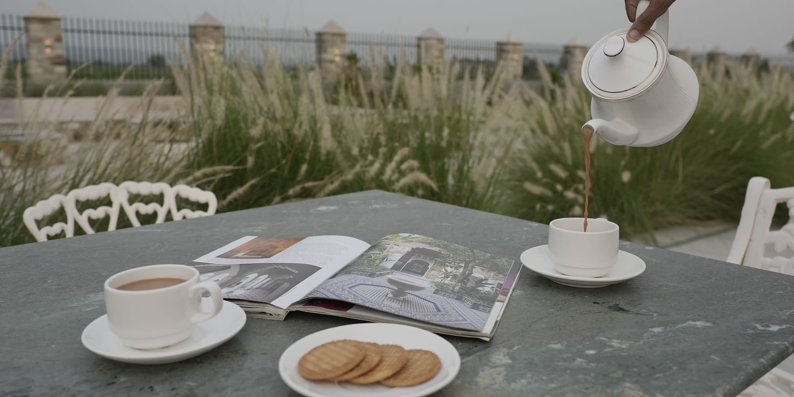 A close-up shot of an outdoor table at Mahendra Niwas with two cups of tea, a plate of biscuits, and a magazine. A person's hand pours tea from a white teapot into one of the cups.