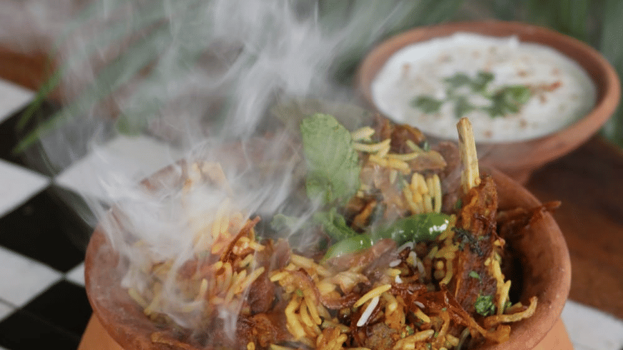 A steaming clay pot filled with biryani, garnished with fried onions and herbs, accompanied by a bowl of raita, beautifully served at Papaya Tree Hotels.