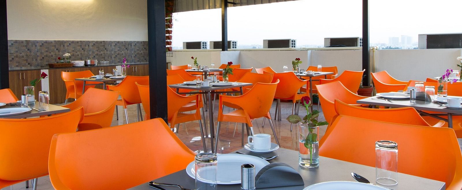 An outdoor dining space under a canopy offers numerous tables with bright orange seating at Sanctum Suites Whitefield, Bangalore.