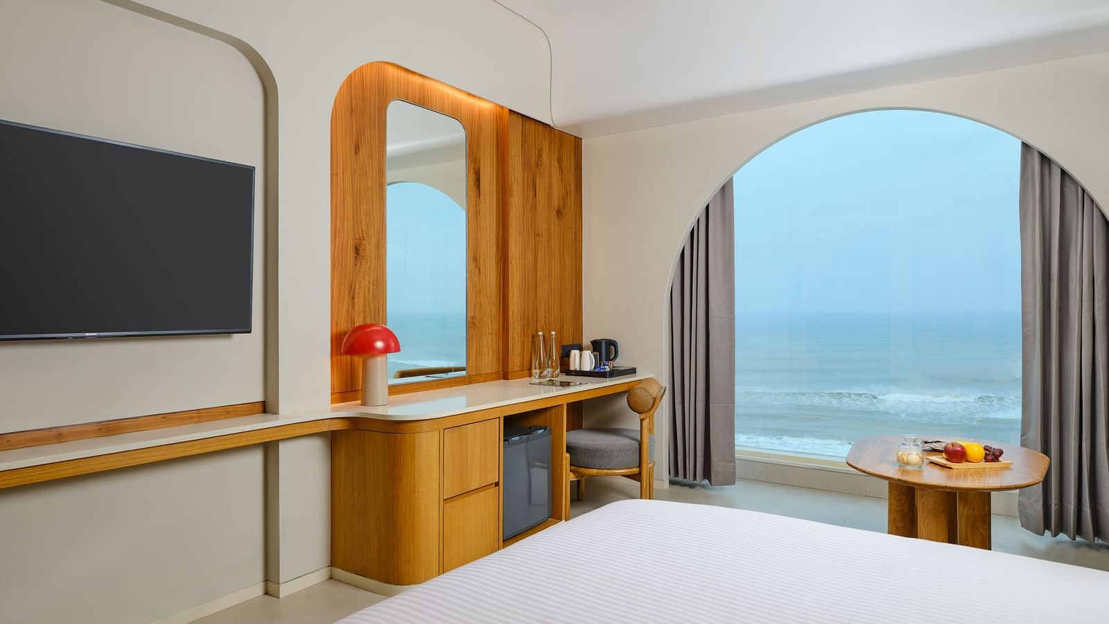 Image featuring a huge TV mounted on the wall next to which a dressing table cum work desk is placed and a huge window with a breathtaking view of the sea
