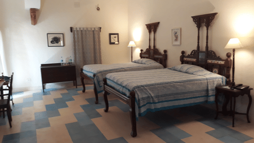 A guest room at Tijara Fort-Palace - 19th Century, Alwar features two beds with striped covers, bedside tables with lamps, chairs, a cabinet, artwork on the walls, and tiled flooring.