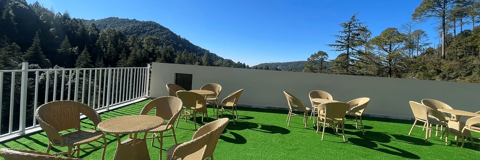 Open terrace area with chairs around tables with a background of trees at DLS Ark Holiday Inn, Banikhet during a sunny day.