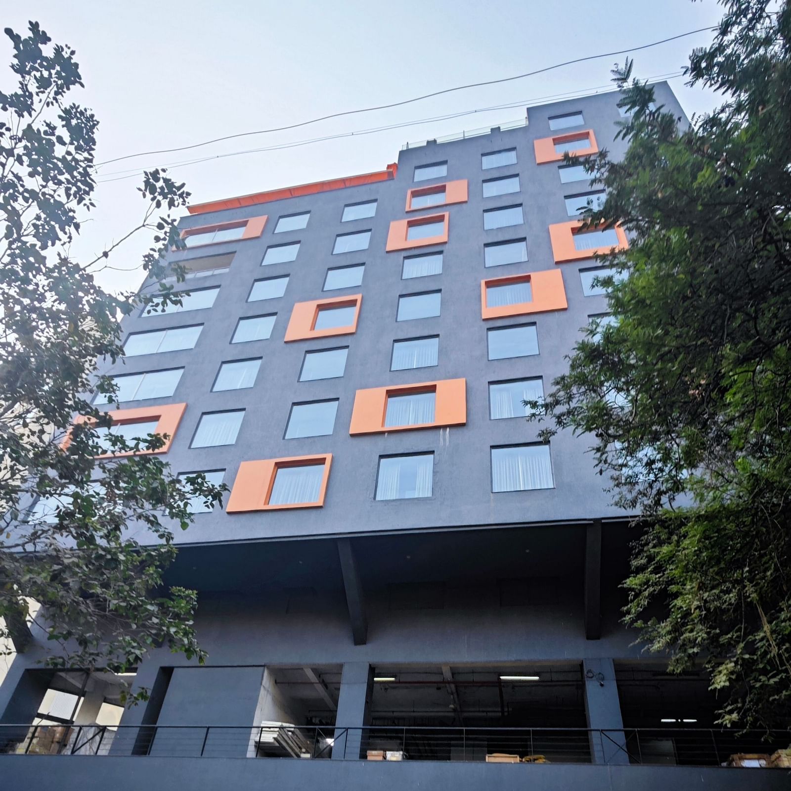 A modern, multi-story hotel building with a dark grey facade and bright orange window accents. The hotel has a covered parking area in front, and there are trees and shrubs surrounding the building.