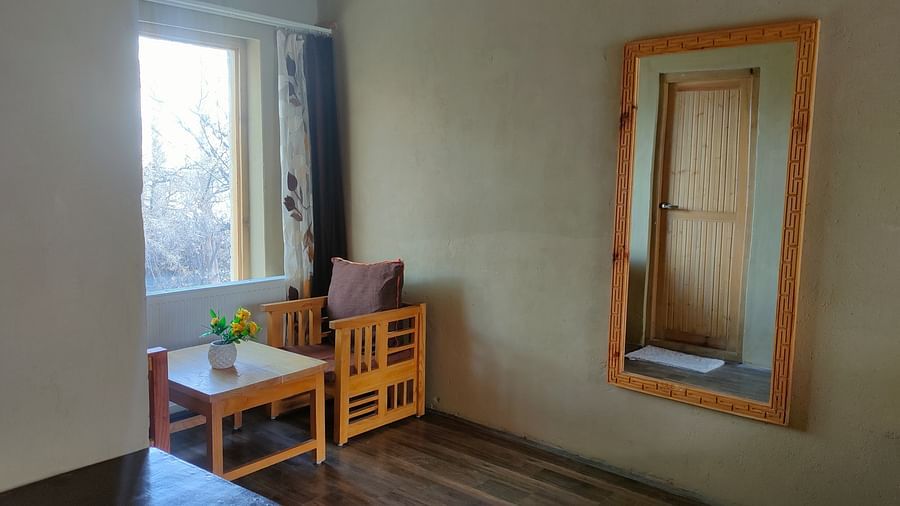A minimalist corner of Deluxe Cottage is captured, featuring a wooden-framed mirror, a small table with a lamp, and a window offering natural light, complemented by a wooden stool at White Sands Resort, Ladakh.