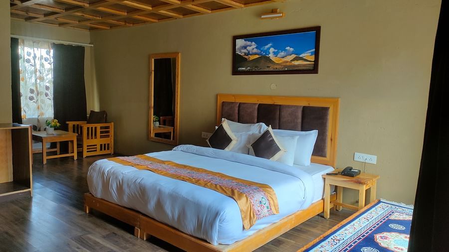 Deluxe Cottage showcases a neatly made king size bed with a striped throw, accompanied by wooden furniture, a decorative ceiling, and a framed picture at White Sands Resort, Ladakh.