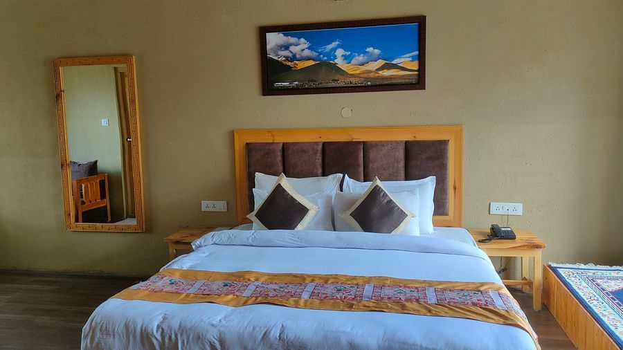 A well-lit Deluxe Cottage features a king size bed with white linens and a patterned throw, flanked by a wooden headboard and bedside table, with a full-length mirror and a framed picture at White Sands Resort, Ladakh.