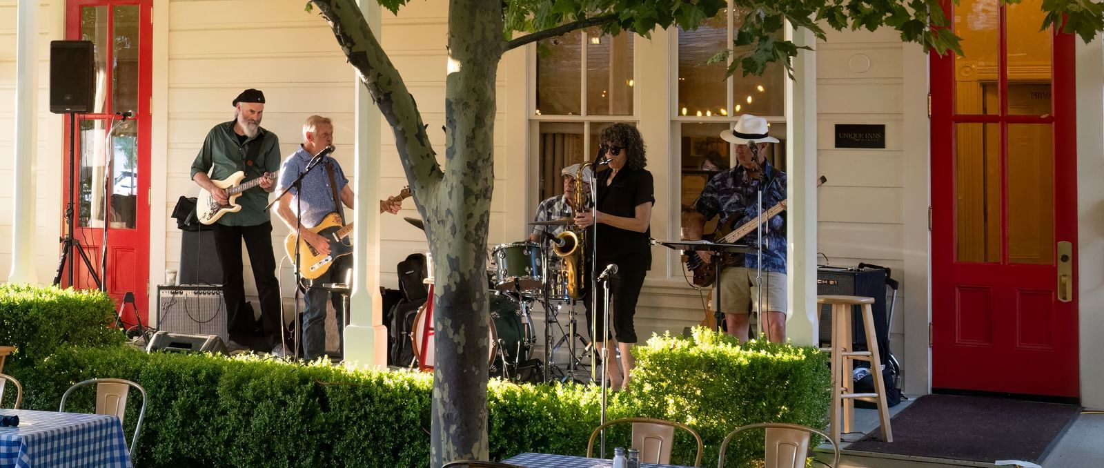 Live jazz musicians perform outdoors under festive string lights, with checkered tables set up at The Tallman Hotel.