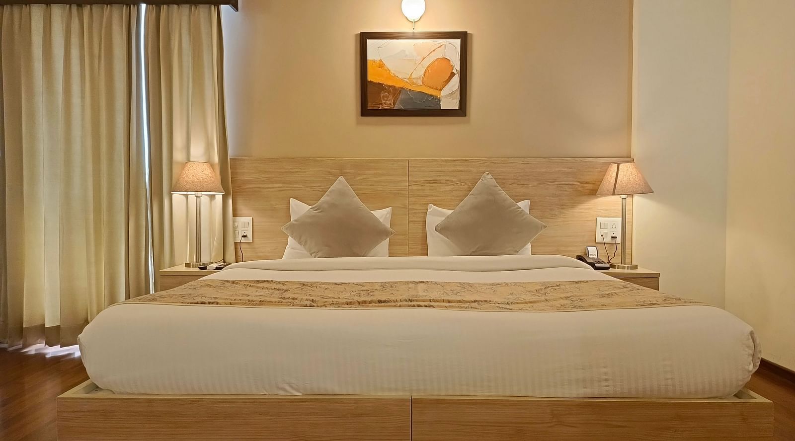 Premium Studio at Starlit Suites, Shirdi features a large double bed with neutral-toned bedding and cushions, bedside tables with lamps, and light-coloured curtains.