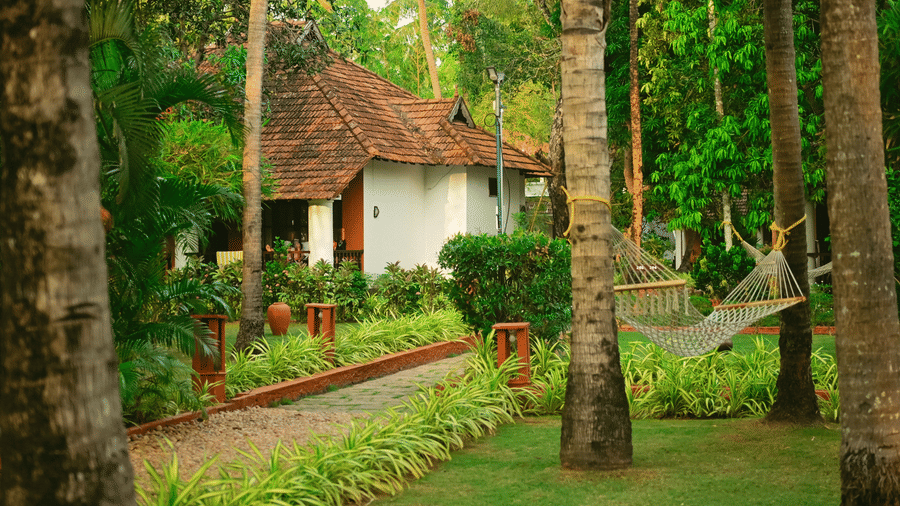 A peaceful garden scene at Ayur On The Beach Nattika featuring a hammock strung between palm trees near a guest cottage.