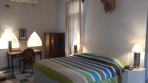 A guest room at Tijara Fort-Palace - 19th Century, Alwar has a bed with striped linens, bedside tables with lamps, a desk with a chair, a wardrobe, an air conditioning unit, and tiled flooring.