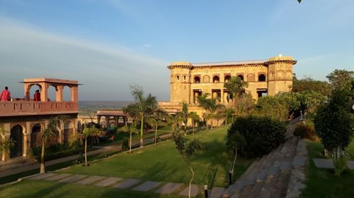 Tijara Fort-Palace - 19th Century, Alwar is pictured with its stone buildings featuring arched openings and turrets, set within green grounds with pathways and trees under a clear sky.