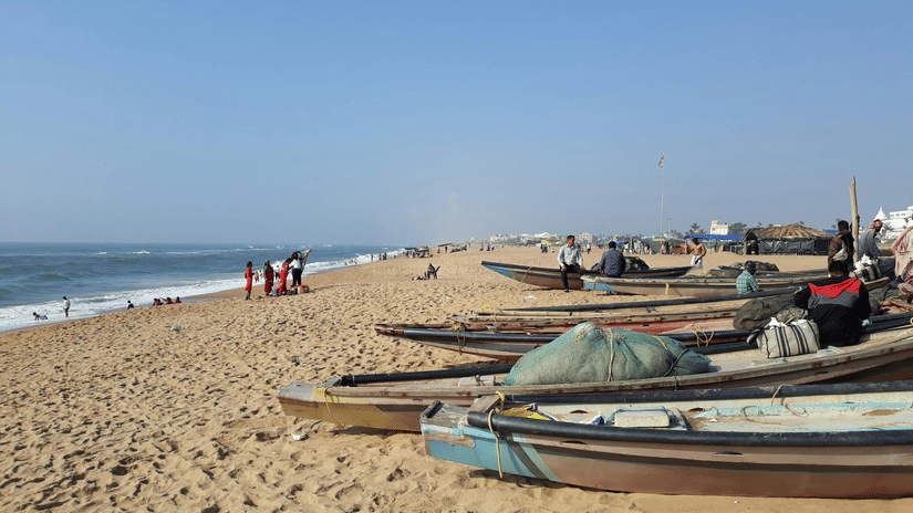 Several small boats rest on a sandy shoreline next to the sea. A few people stand in the distance along the water.