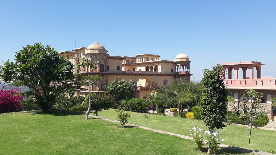 Tijara Fort-Palace - 19th Century, Alwar is shown with its stone architecture, arched openings, domes, surrounding green lawns, trees, and a clear sky.