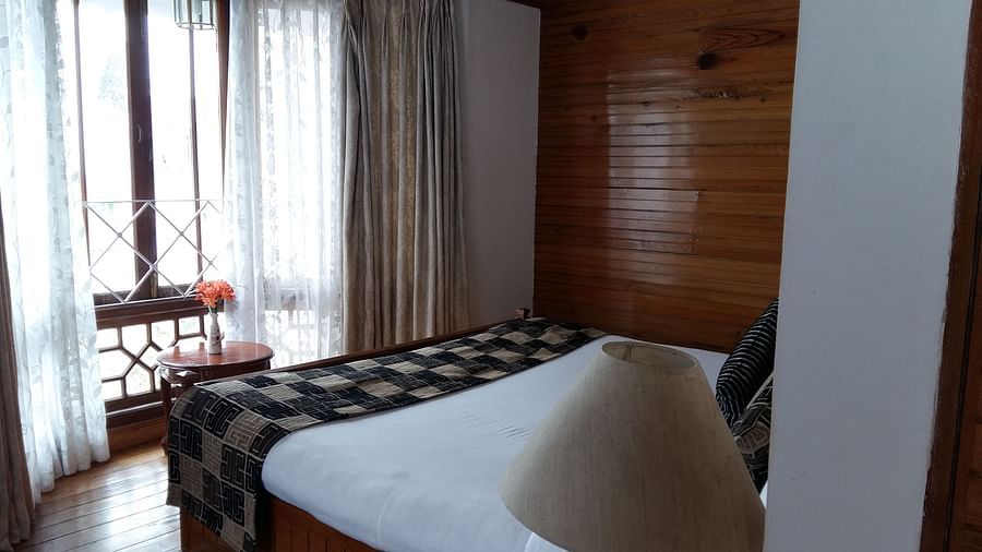 The Executive Suite at Cedar Inn in Darjeeling features a double bed with a bedside table, lamps facing a glass curtained window.