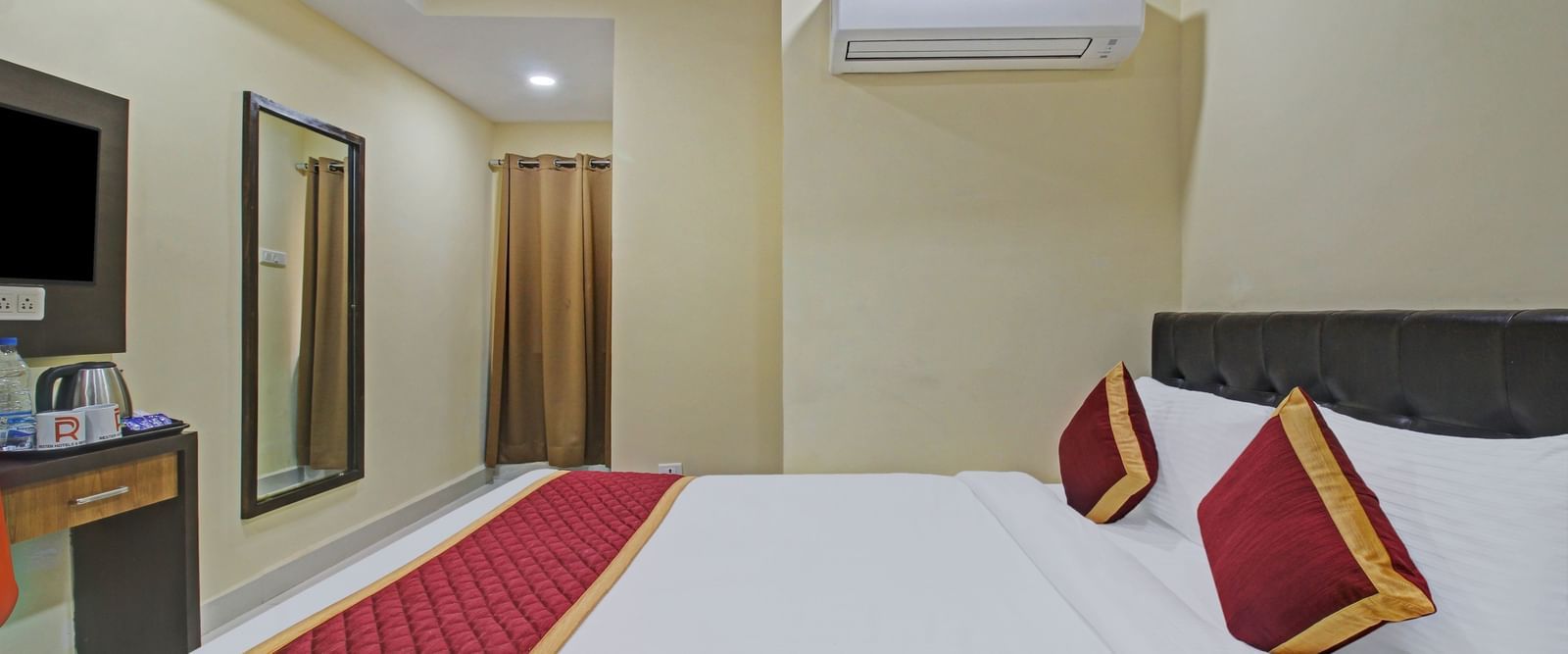 A bedroom with a double bed featuring white linens and red accent pillows, a black upholstered headboard, facing the TV mounted on the wall next to a mirror at Lemonridge Hotels, Kukatpally