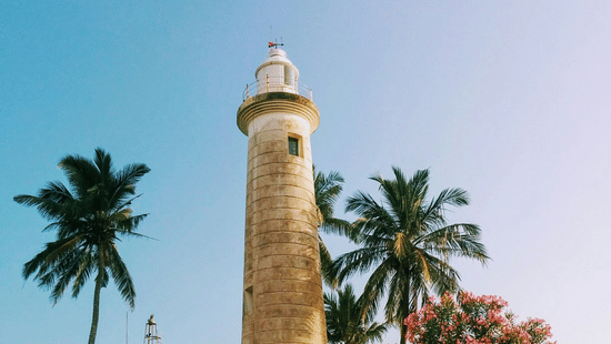 Daytime view of the Galle Dutch Fort lighthouse in Sri Lanka with white walls, palm trees, and a bright blue sky.