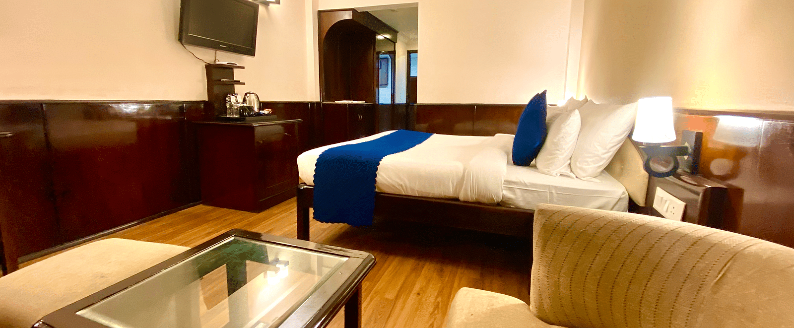 A view from a cushioned chair showcasing the bed and wall-mounted television at DLS Park Grace, Haridwar.