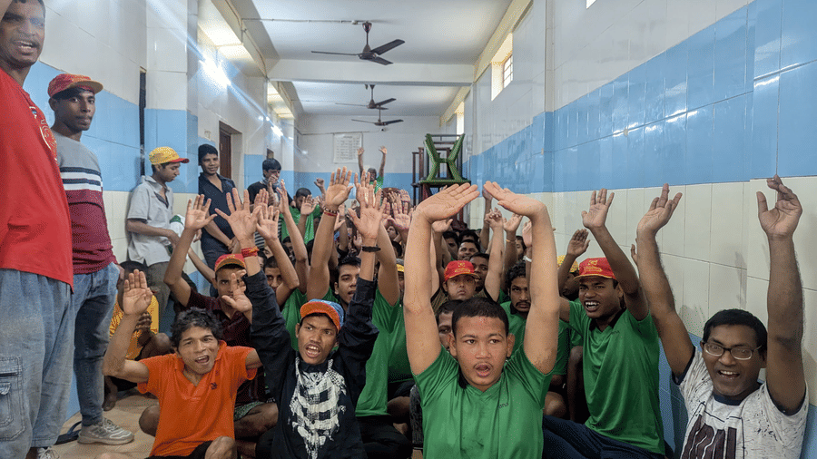 A group of children and young adults with special needs sit indoors, smiling and raising their hands enthusiastically during a group session.