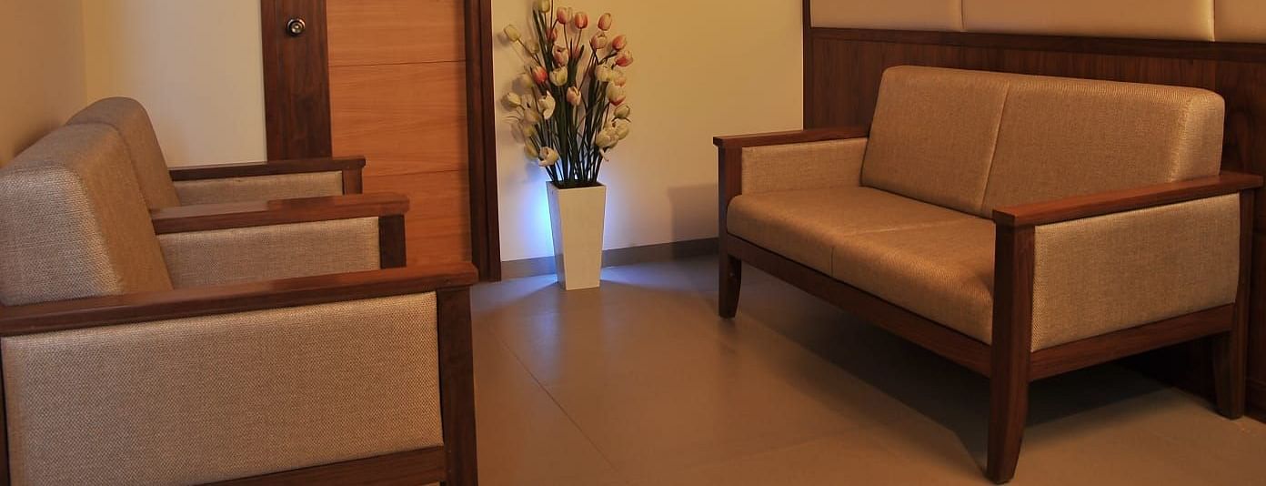 Suite's seating area at Dhanalakshmi Srinivasan Hotels, Perambalur shows two light brown couches with wooden frames facing each other, a wooden door, and a white vase with artificial tulip flowers.