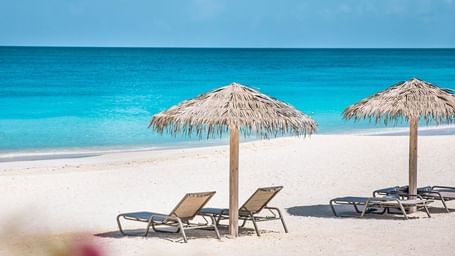 A pristine white sand beach with two thatched umbrellas and lounge chairs, facing a brilliant turquoise ocean.