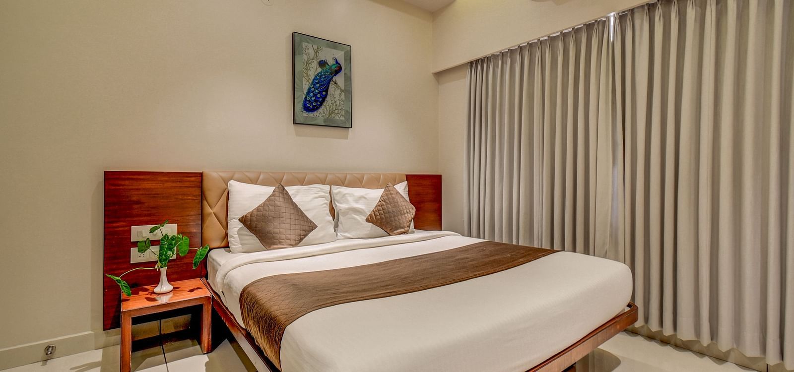 A spacious room at Harigovinda's JNStay with a large bed, a bedside table, large windows fully covered with curtains, and a wall-mounted photo frame above the headboard