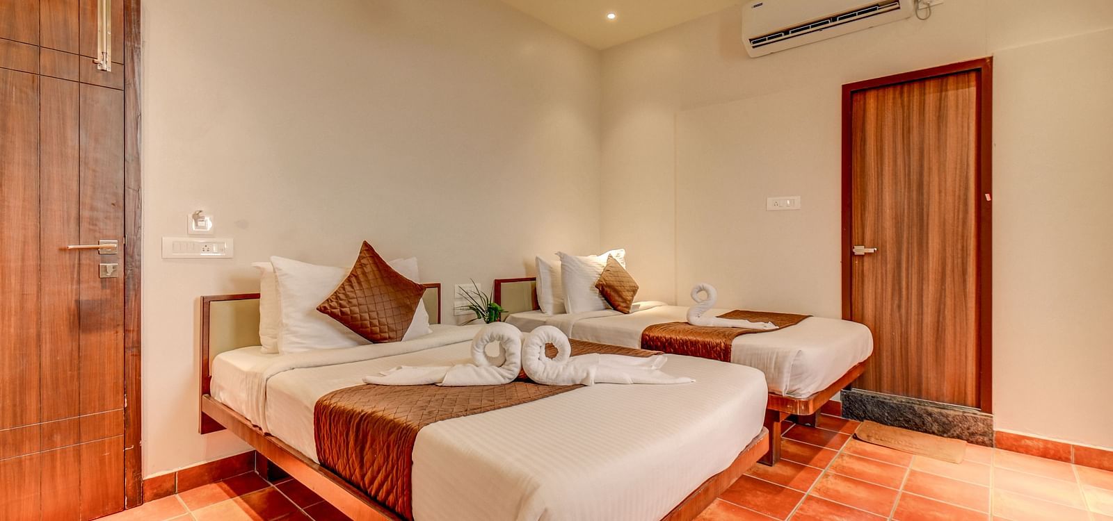 Two neatly made beds placed side by side, with doors on both sides and a wall-mounted AC above one door at Harigovinda's JNStay