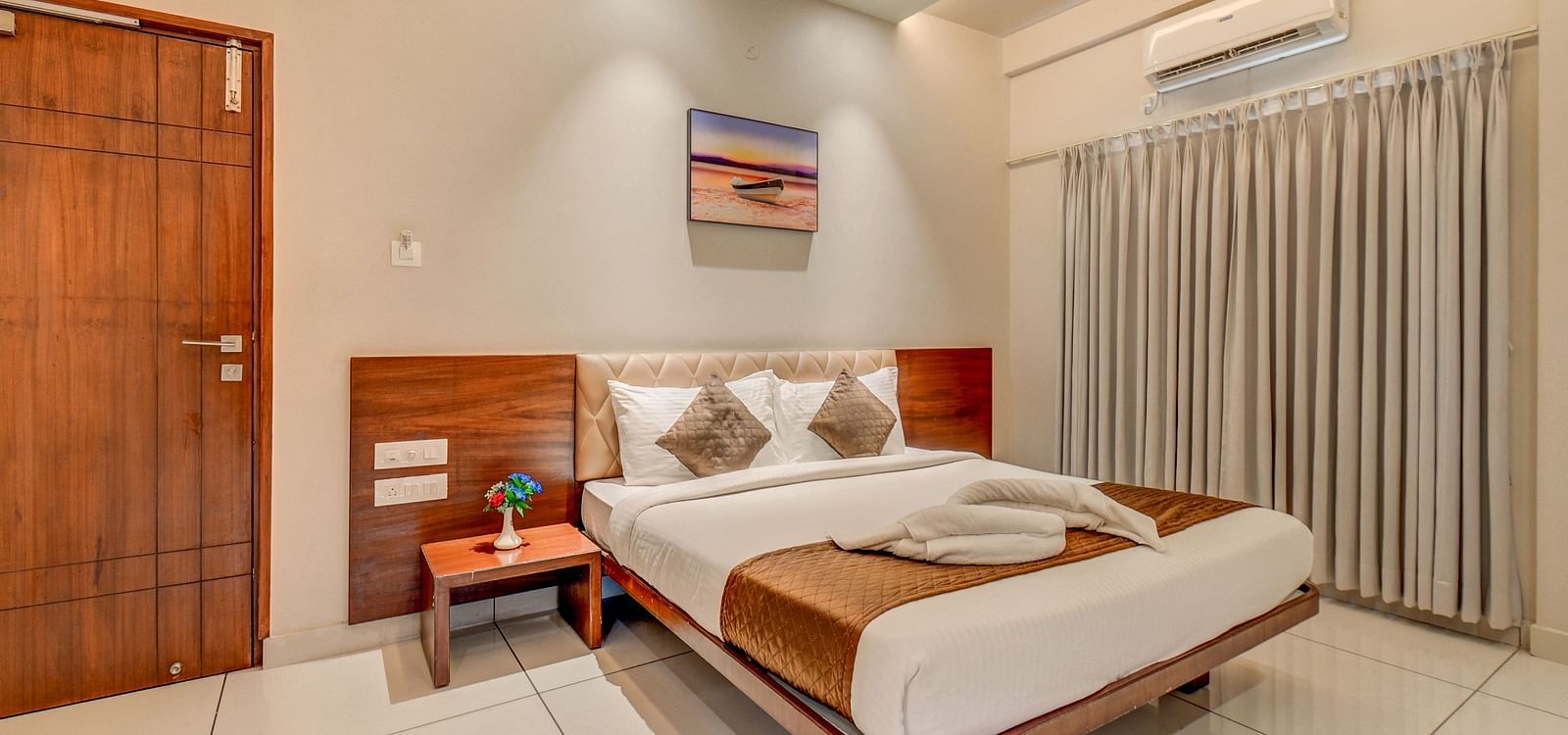 A spacious room at Harigovinda's JNStay with a large bed, wooden headboard, bedside table with a plant, a door, and curtains covering the window