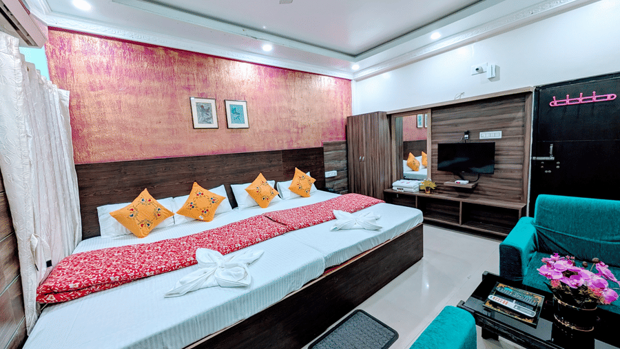 A modern hotel bedroom with a large double bed, bright orange pillows, a television unit, and turquoise armchairs.