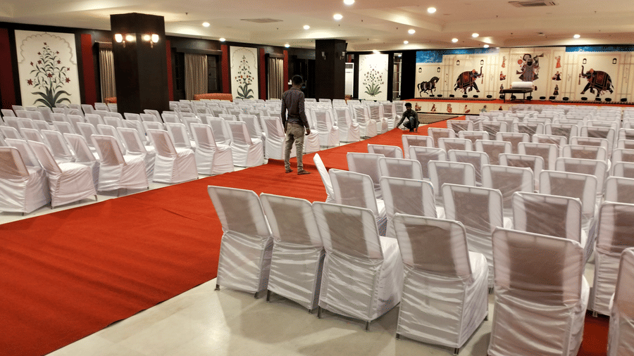 An indoor Raj Mahal banquet hall in Nathdwara at Shri Vallabh Vilas Lords Plaza, decorated with white chairs in evenly spaced rows and a red carpet aisle leading to the stage, featuring two people assisting with decorations.