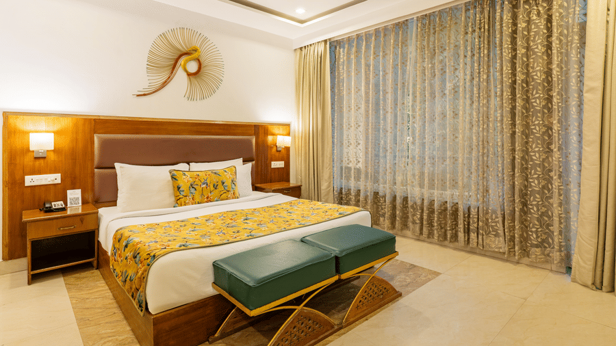 A room featuring a wooden bed, 2 bedside tables, 2 vanity stools, a decorative wall piece, and full-length curtains | Summit by the Ganges Beach Resort & Spa, Rishikesh